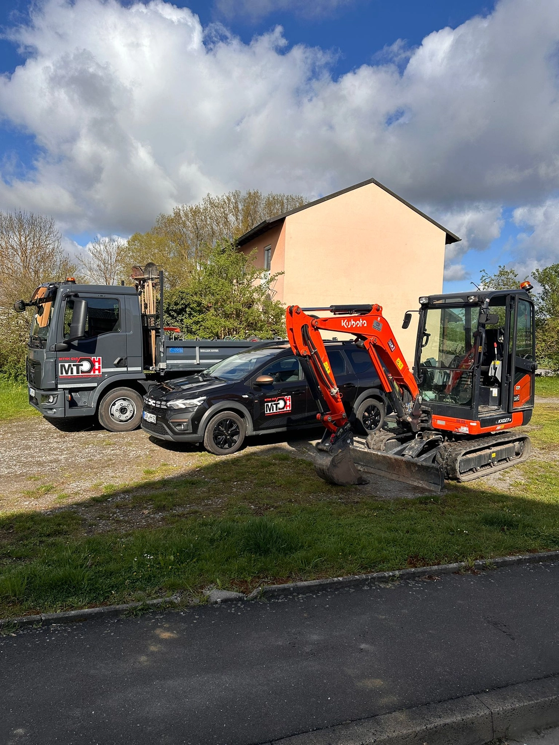 MTHH GbR - Kabeltragsystembau, Regalbau/Prüfung und Tiefbau - Ein orangefarbener Kubota-Kompaktbagger, ein schwarzer Geländewagen und ein grauer Muldenkipper parkten zusammen auf einer Schotterfläche vor einem Haus.