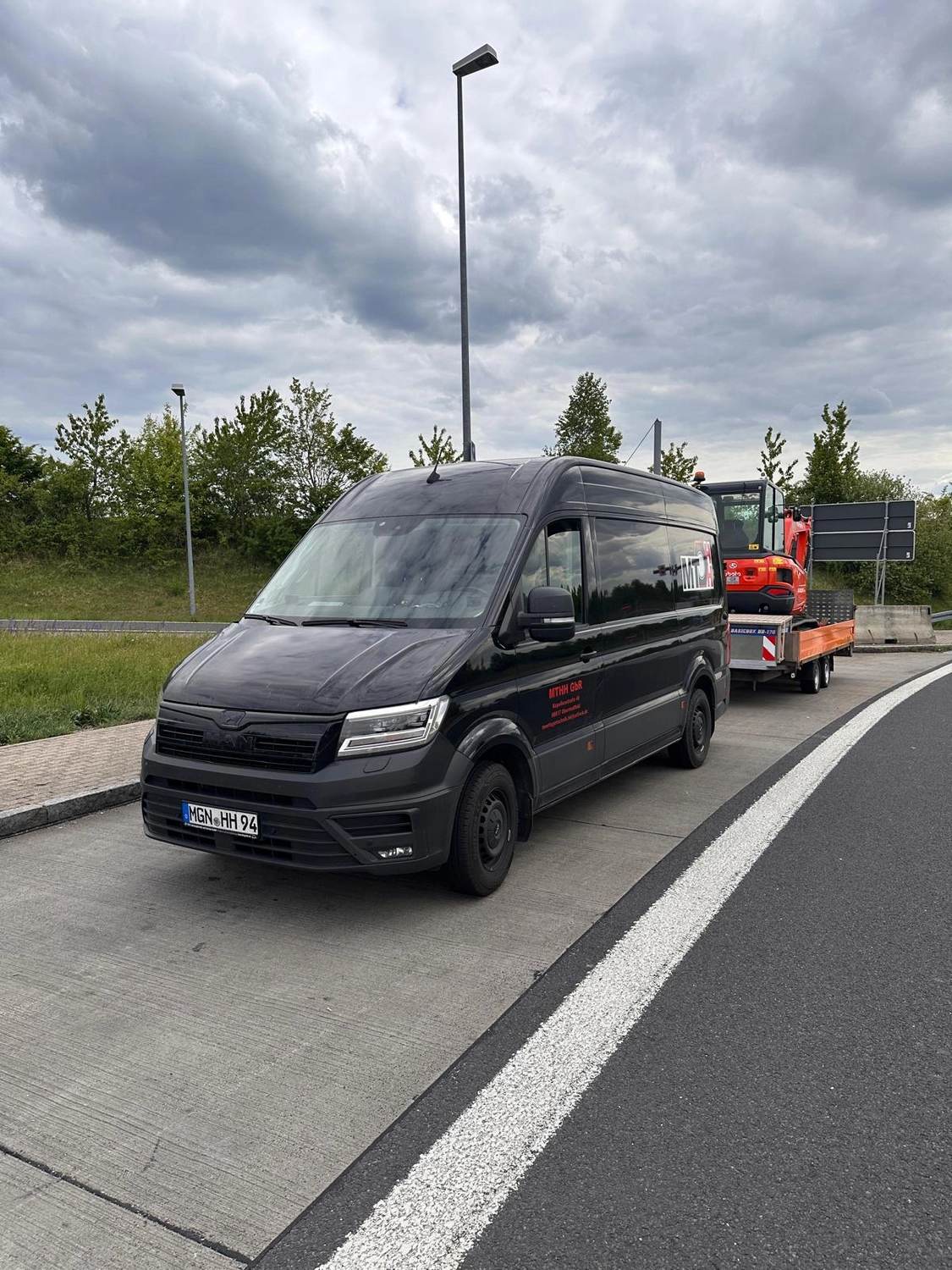 MTHH GbR - Kabeltragsystembau, Regalbau/Prüfung und Tiefbau - Ein schwarzer MAN TGE Transporter parkte am Straßenrand und zog unter bewölktem Himmel einen kleinen orangefarbenen Bagger auf einem Anhänger.