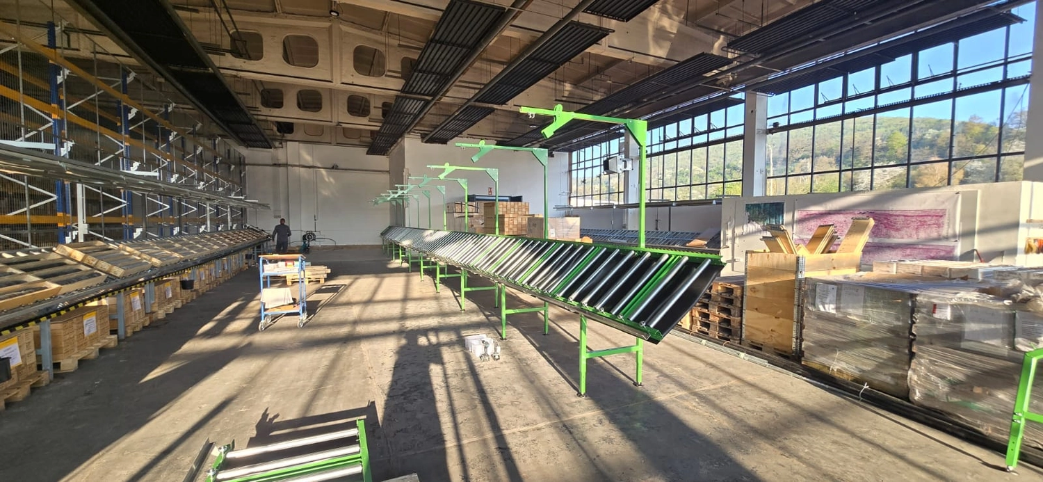 MTHH GbR - Kabeltragsystembau, Regalbau/Prüfung und Tiefbau - Eine Weitwinkelaufnahme auf Augenhöhe zeigt eine helle, geräumige Lagerhalle mit hohen Decken und großen Fenstern auf der rechten Seite, durch die Bäume und ein Hügel zu sehen sind. Zwei lange Reihen grün umrahmter Rollenförderer erstrecken sich in die Ferne; am Ende der linken Reihe steht eine Person. Links davon befinden sich hohe Industrieregale, rechts sind Holzpaletten und -kisten in Plastikfolie verpackt. Sonnenlicht strömt durch die Fenster und wirft lange Schatten auf den Betonboden.